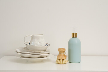  Bottle of dishwashing detergent with a natural brush and a stack of dishes on a minimalist white surface creates a perfect aesthetic