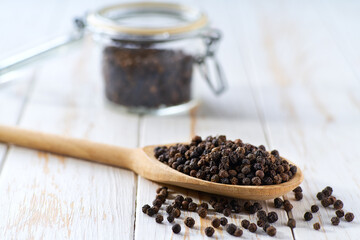 black pepper seeds in a wooden scoop and in glass storage jar on a white wooden table, selective focus.