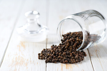 black peppercorns spill out of a glass storage jar on a white wooden table, selective focus.