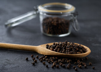 black pepper seeds in a wooden scoop and in glass storage jar on a black stone table, selective focus.