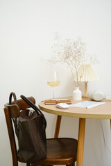 A calm and serene workspace showcasing a beautiful wooden table, stylish decorative elements, and a glass of fine wine, keyboard, books in a basket, headphones, bag on the shair.