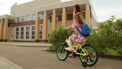 Young caring mother help little daughter pupil with backpack riding on bike to elementary school outdoor. Happy family mom and female kid child driving cycle transportation at schoolyard campus road