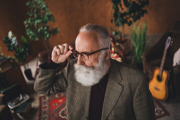 Sophisticated gentleman with gray hair and beard adjusting glasses, surrounded by a vintage interior including a guitar and plants