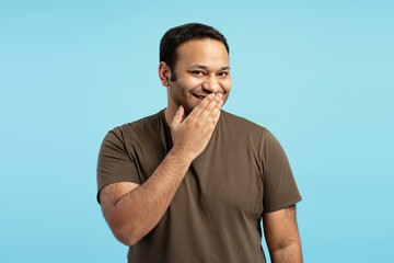 Indian man covering mouth with hand while smiling on blue background
