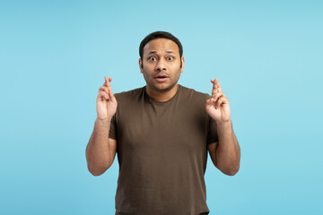 Fototapeta premium Hopeful man crossing fingers wishing for good luck on blue background