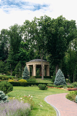 Classical style rotunda with columns and domed roof standing in a well-maintained park with manicured lawn, bushes, and trees, enjoying the sunlight of a beautiful summer day