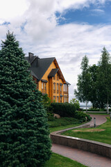 Modern wooden cottage with dark roof and chimneys surrounded by evergreen trees and bushes under cloudy sky in countryside during summer day