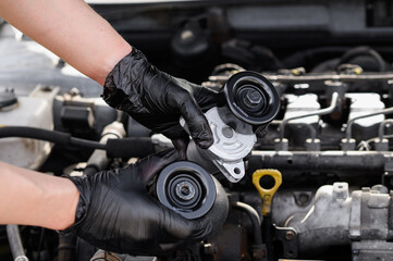 Fototapeta premium Auto mechanic repairing under hood of car, holding belt pulley and serpentine belt tensione against background of car engine compartment, close-up. Auto parts replacement, car maintenance, autoservice
