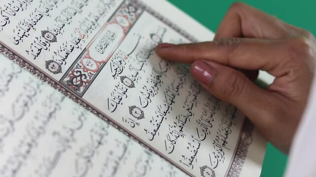 A hand points at an open Quran with Arabic scrip, an Islamic religious text, close up. The page is displayed with arabic calligraphy and decorative ornaments isolated on a green background or green sc