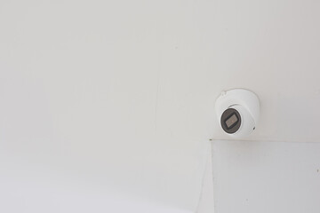 white dome security camera mounted on a white ceiling. The camera has a black lens area and is...