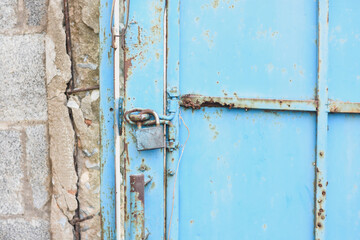 close-up view of a blue metal door with significant rust and corrosion. The door is secured with a rusty padlock and has a metal latch.