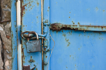 close-up view of a blue metal door with significant rust and corrosion. The door is secured with a rusty padlock and has a metal latch.