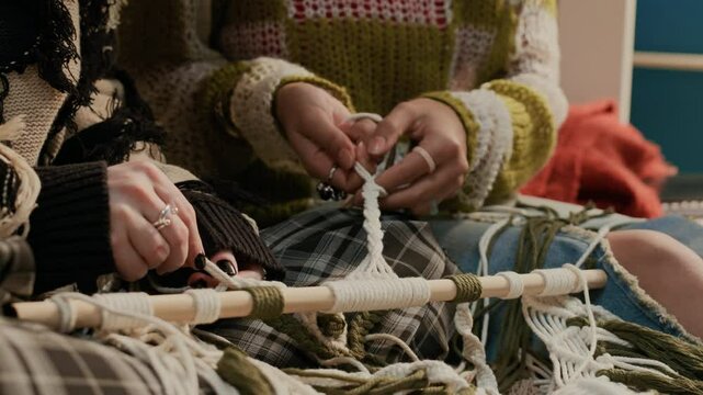 Close-up view of two unknown young women dressed in sweaters weaving interior piece using macrame technique