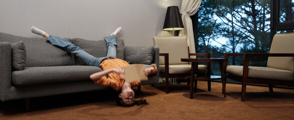 A young woman joyfully relaxing upside down on a couch in a cozy living room, expressing carefree...
