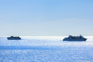 Obraz premium Two ships sailing on a calm, shimmering blue sea under a clear sky. Larger ship is on the right side of the image, while the smaller ship is on left, both leaving trails in water as they move forward