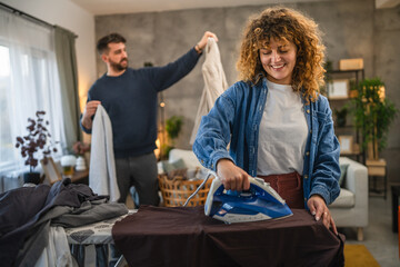 A woman iron shirts on an ironing board, while a man folds up them