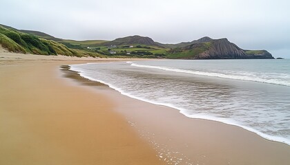 Sandy beach meets ocean waves under a cloudy sky, with green hills and distant houses in the background.