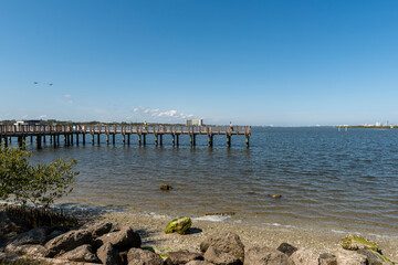 A fishing pier juts out into the Halifax River seen from the rocky river bank of a Port Orange, Volusia county park, Florida.

