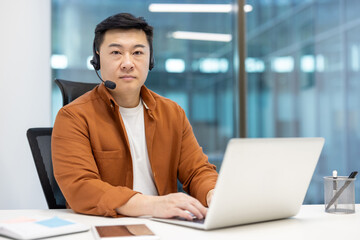 Portrait of an Asian worker with a headset and a laptop inside the office. The man is looking at...