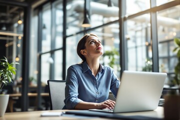 Woman in office with thoughtful expression, working on laptop in modern workspace with natural light and greenery.