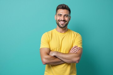 Fototapeta premium Smiling man in yellow shirt standing with arms crossed on teal background.