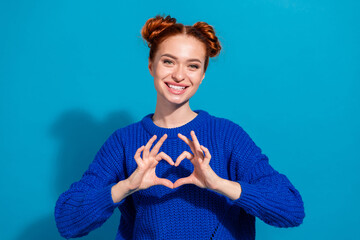 Young red-haired woman forming heart gesture with hands against blue background in stylish sweater