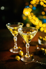 Two martini glasses with green olives stand on a dark table with scattered olives, while blurred golden lights shimmer in the background, creating a festive and elegant atmosphere.