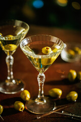 Two martini glasses filled with clear liquid and garnished with green olives, placed on a stylish bar setting with golden bokeh lights in the background.
