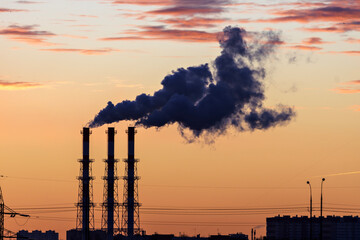 Factory chimneys release dense dark smoke into the air, colored by sunset hues. Industrial pollution of a metropolis.