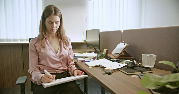 A young businesswoman sits at her desk in a modern office, focused on writing in her notepad. Natural light filters through the blinds, creating a calm ambiance.