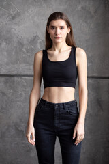 A professional modeling snapshot of a young female model in a black crop top and high-waisted jeans, posing against a textured gray wall. Capturing her natural elegance, toned physique, and confident 