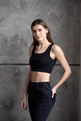 Fototapeta premium A professional modeling snapshot of a young female model in a black crop top and high-waisted jeans, posing against a textured gray wall. Capturing her natural elegance, toned physique, and confident 