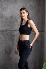Fototapeta premium A professional modeling snapshot of a young female model in a black crop top and high-waisted jeans, posing against a textured gray wall. Capturing her natural elegance, toned physique, and confident 