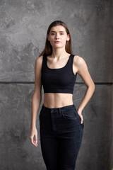 Fototapeta premium A professional modeling snapshot of a young female model in a black crop top and high-waisted jeans, posing against a textured gray wall. Capturing her natural elegance, toned physique, and confident 