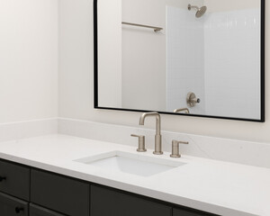 A bathroom sink detail with a brushed nickel faucet on a white marble countertop, dark vanity cabinet, and a white tiled shower seen through the black frame mirror.