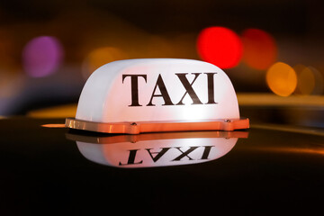A brightly lit taxi sign rests on top of a black vehicle, reflecting the glow of city lights in the evening. The urban atmosphere creates a vibrant backdrop.