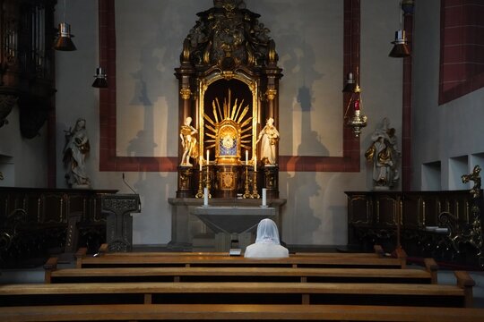 A lady wearing a white jacket and white lace veil sits in prayer in front of the altar of a church