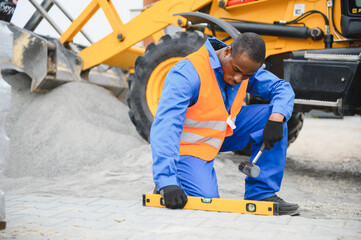 African american master in gloves lays paving stones in layers. Garden brick pathway paving by professional paver worker. Laying gray concrete paving slabs in house courtyard on sand foundation base