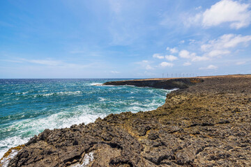 Waves crashing against rugged volcanic coastline of Arikok National Park in Aruba, with wind turbines standing along horizon under bright blue sky.