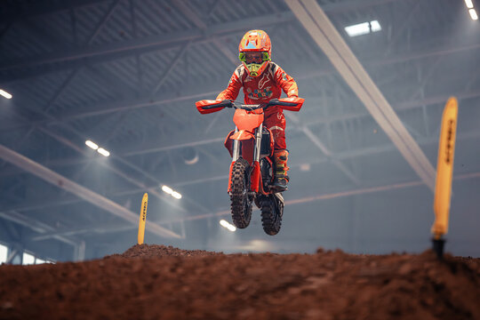  a person wearing a helmet, goggles, gloves, and shoes, riding a dirt bike in the air on top of a dirt field In the background, there are rods and lights on the cei