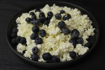 The cottage cheese in the black plate. On a black background. Close up of cottage cheese with blueberries. Honey watering curd on black background. 