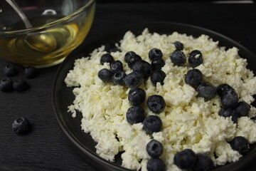 The cottage cheese in the black plate. On a black background. Close up of cottage cheese with blueberries. Honey watering curd on black background. 