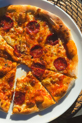 Pepperoni pizza on a white plate, placed on a woven placemat. Pizza has golden-brown crust, melted cheese, and pepperoni slices. One slice partially removed, revealing gooey cheese and toppings