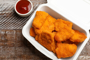 Delicious chicken nuggets in takeaway container and ketchup on wooden table, above view