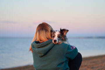 A person walks along the beach with a Border Collie as the sun sets, casting a soft glow. The pair enjoys a tranquil and scenic moment by the water.