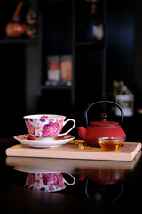 Floral teacup and red teapot set on a wooden board reflecting in a dark surface