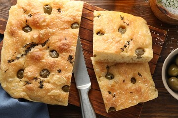 Pieces of delicious focaccia bread with olives, thyme, salt and knife on wooden table, flat lay