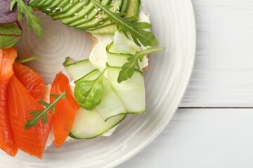 Delicious bagel with salmon, cream cheese, cucumber and avocado on white wooden table, top view