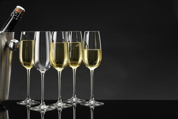 Bottle of sparkling wine in cooler bucket and glasses on black mirror surface. Space for text