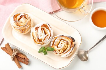 Freshly baked apple roses with mint, honey, cinnamon sticks and tea on white table, flat lay